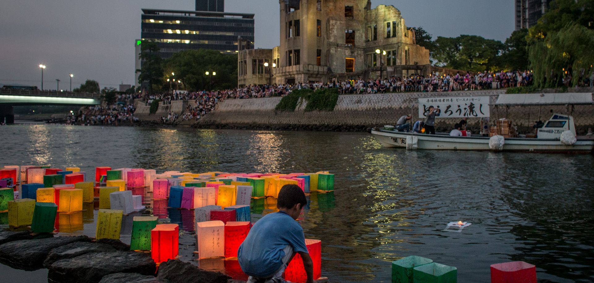 A boy floats a candle lit paper lantern on the river in front of the Atomic Bomb Dome.