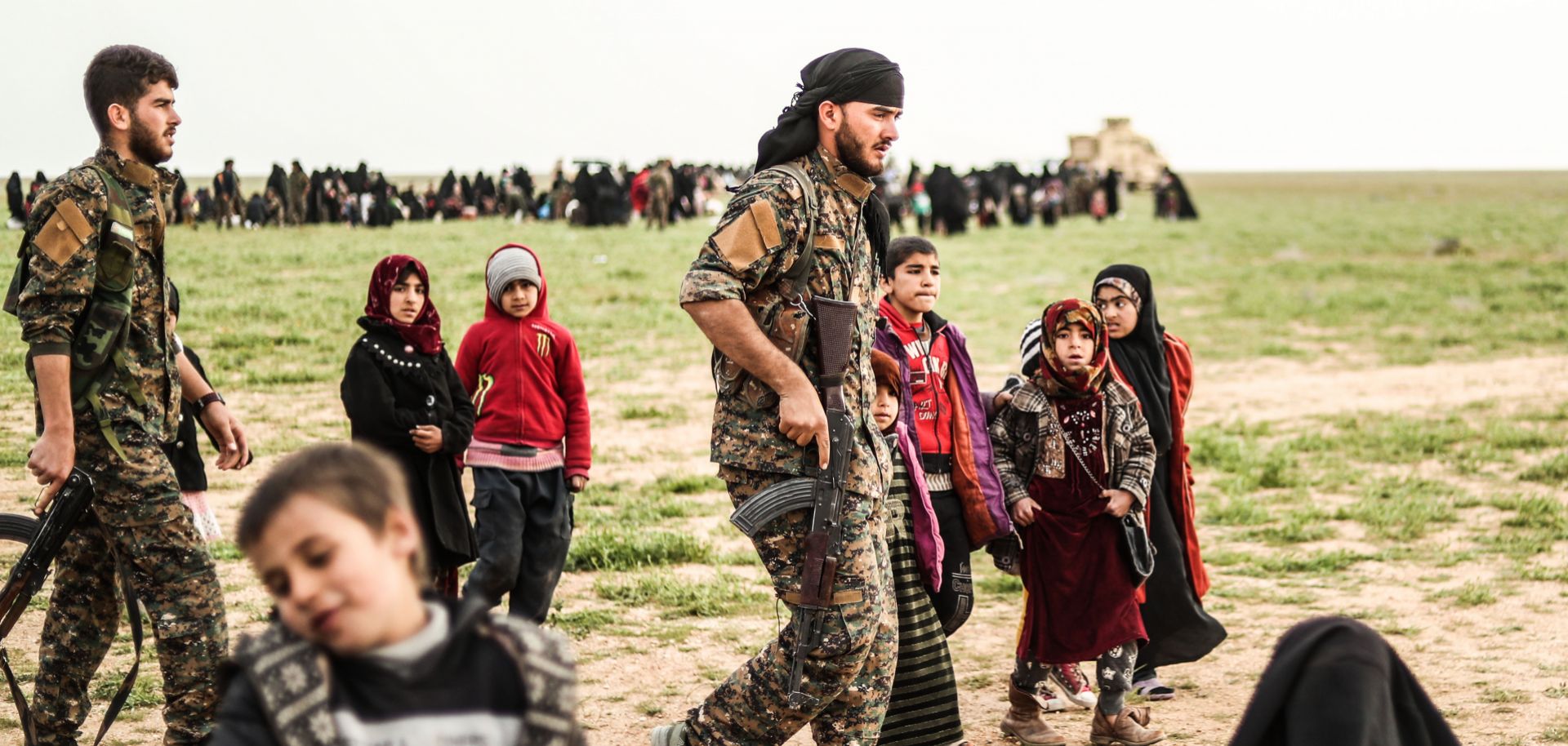 Fighters with the Syrian Democratic Forces walk past civilians at a screening area for evacuees from the Islamic State's embattled holdout in Baghouz, Syria, on Feb. 26, 2019. 